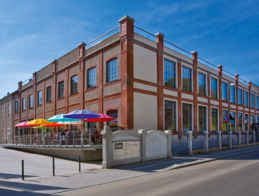 Außenansicht des Textilmuseums Augsburg: Historischer roter Backsteinbau mit hohen Rundbogenfenstern, ergänzt durch einen modernen gläsernen Eingangsanbau, vor klarem Himmel